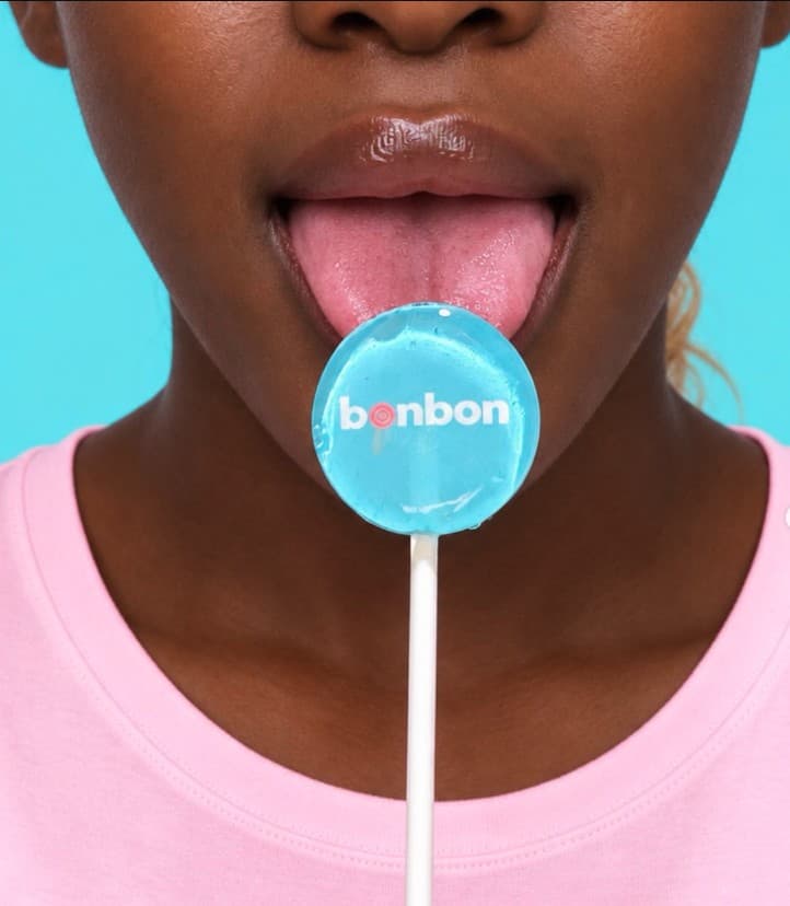 A blue bonbon lollipop with the bonbon logo set inside the sugar, held to a tongue against turquoise.